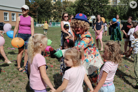Klaun w niebieskim meloniku rozmawia przez mikrofon z dziećmi. Trzy dziewczynki trzymają się za ręce i rozmawiają z klaunem.