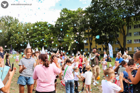 Dzieci i dorośli bawią się i tańczą na przeciwko dmuchanego zamku. W powietrzu unosi się kolorowe confetti.