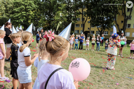 Dzieci ustawione są w kręgu. Na głowach mają srebrne czapeczki urodzinowe. W środku stoi klaun w niebieskim meloniku i przemawia do dzieci.