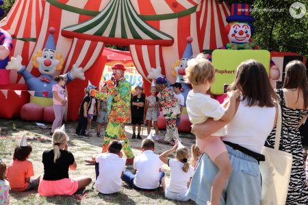 Klaun w czerwonym meloniku rozmawia z dziećmi przez mikrofon. Przed nim stoi duża grupa dzieci.