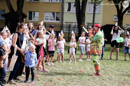 Klaun w czerwonym meloniku rozmawia z dziećmi przez mikrofon. Przed nim stoi duża grupa dzieci.