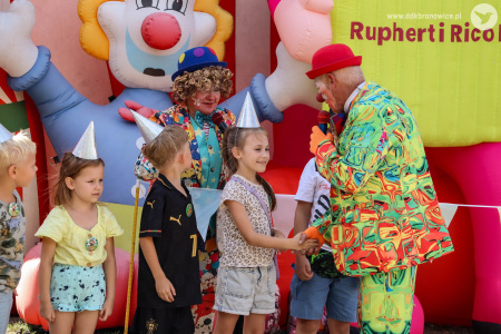 Klaun w czerwonym meloniku rozmawia z grupką dzieci. Dzieci mają nałożone srebrne czapeczki.