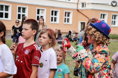 Klaun w niebieskim meloniku farbuje włosy dzieciom. Dzieci ustawione są w rzędzie.