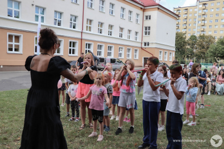 Dzieci gwizdają w papierowe gwizdki przed Sylvettą Księżniczką Wiatru. W tle rodzice siedzą na czarnych krzesełkach.
