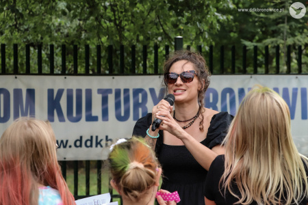 Sylvetta śpiewa do mikrofonu. Przed nią bawi się grupka dzieci. Artystka ubrana jest w czarna sukienkę.