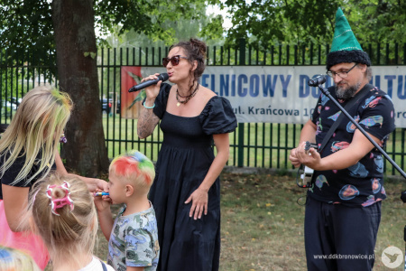 Sylvetta śpiewa do mikrofonu. Przed nią bawi się grupka dzieci. Artystka ubrana jest w czarna sukienkę. Obok niej stoi gitarzysta w zielonej długiej czapce.