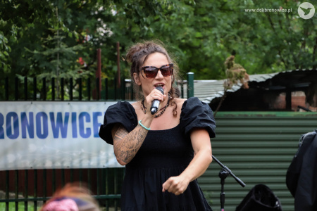 Sylvetta śpiewa do mikrofonu. Przed nią bawi się grupka dzieci. Artystka ubrana jest w czarna sukienkę.
