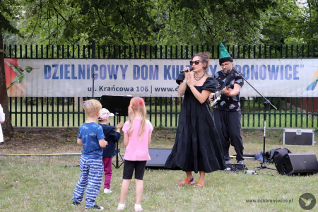 Sylvetta śpiewa do mikrofonu. Przed nią bawi się grupka dzieci. Artystka ubrana jest w czarna sukienkę. Obok niej stoi gitarzysta w zielonej długiej czapce.