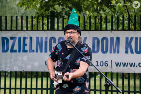 Mężczyzna w zielonej czapce gra na gitarze. śpiewa do mikrofonu. Za nim biały baner Dzielnicowego Domu Kultury Bronowice.