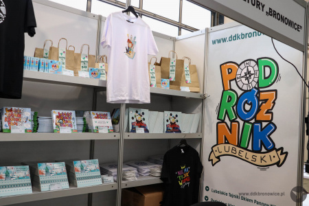 Białe stoisko Dzielnicowego Domu Kultury Bronowice. Na półkach książki, koszulki i rozwieszone plakaty.