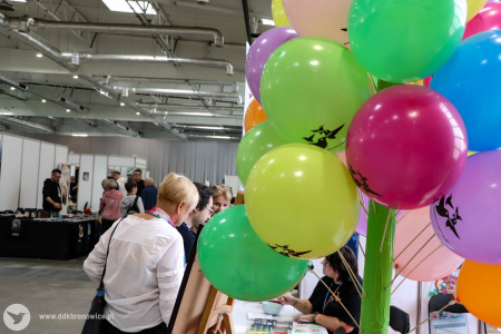 Po prawej stronie pęk kolorowych balonów Dzielnicowego Domu Kultury Bronowice. Na balonach widać białe kolibry. Po lewej stronie osoba ogląda produkty wystawione na stosiku.