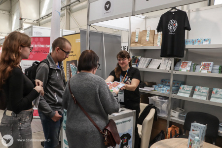 Na stoisku Dzielnicowego Domu Kultury Bronowice trzy osoby kupują książki. W tle książki i koszulki.