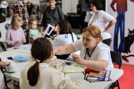 Na stoisku warsztatowym dzieci i dorośli pracują nad swoimi artystycznymi zakładkami do książek i pocztówkami. Pomocą służą pracowniczki DDK Bronowice w białych koszulkach.
