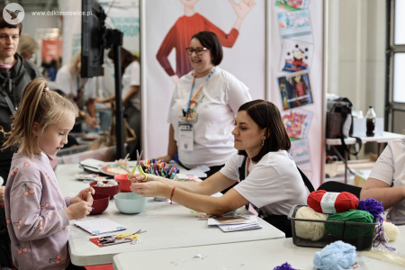 Na stoisku warsztatowym dzieci i dorośli pracują nad swoimi artystycznymi zakładkami do książek i pocztówkami. Pomocą służą pracowniczki DDK Bronowice w białych koszulkach.