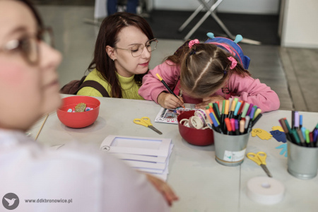 Na stoisku warsztatowym dzieci i dorośli pracują nad swoimi artystycznymi zakładkami do książek i pocztówkami. Pomocą służą pracowniczki DDK Bronowice w białych koszulkach.