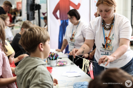 Na stoisku warsztatowym dzieci i dorośli pracują nad swoimi artystycznymi zakładkami do książek i pocztówkami. Pomocą służą pracowniczki DDK Bronowice w białych koszulkach.