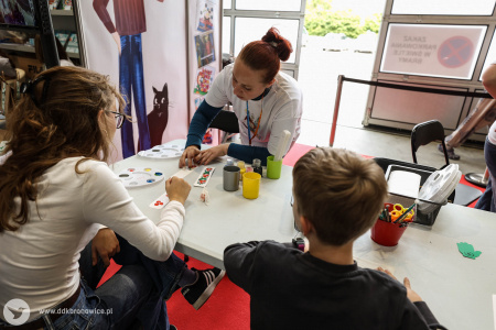 Na stoisku warsztatowym dzieci i dorośli pracują nad swoimi artystycznymi zakładkami do książek i pocztówkami. Pomocą służą pracowniczki DDK Bronowice w białych koszulkach.