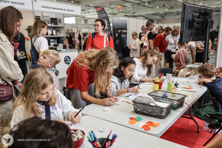 Na stoisku warsztatowym dzieci i dorośli pracują nad swoimi artystycznymi zakładkami do książek i pocztówkami. Pomocą służą pracowniczki DDK Bronowice w białych koszulkach.