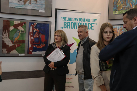 Obok zawieszonych obrazów stoi dyrektor Dzielnicowego Domu Kultury Bronowice pani Aleksandra Szulc0Choma. Za nią stoi plakat Dzielnicowego Domu Kultury.