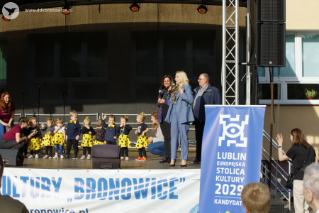 Święto Dzielnicy Dziesiąta. Na scenie elegancko ubrani prowadzący zapowiadają kolejne występy. W tle sceny widać szykujące się dzieci.