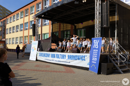 Święto Dzielnicy Dziesiąta. Na scenie elegancko ubrani prowadzący zapowiadają kolejne występy. W tle sceny widać szykujące się dzieci.