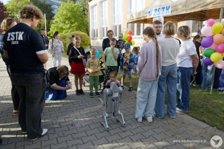 Przy wejściu do szkoły stoi grupka dzieci. Chłopiec w czarnej koszulce kieruje zdalnie małego, szarego robota. Osoby dorosłe robią zdjęcia.