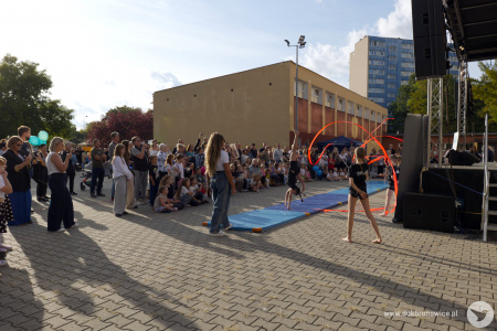 Przed sceną występ gimnastyki artystycznej. Dziewczynki używają kolorowych szarf jedna z nich stoi na rękach na niebieskiej macie.