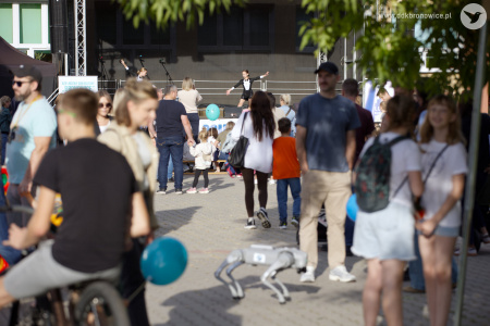 W tle, na scenie występ dwóch tancerek. Na pierwszym planie, tłum ludzi. Po chodniku idzie robot w kształcie psa.