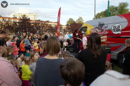 Duża publiczność zebrała się by oglądać występ magika. Magik ma czerwoną marynarkę. Obok niego stoi chłopiec. Za nimi stoi duży czerwony samochód.