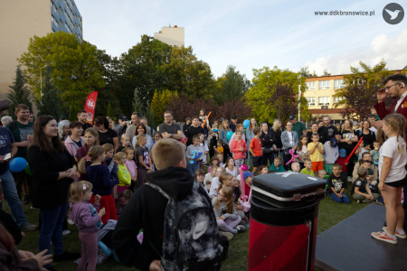 Duża publiczność zebrała się by oglądać występ magika. Magik ma czerwoną marynarkę. Obok niego stoi chłopiec. Za nimi stoi duży czerwony samochód.