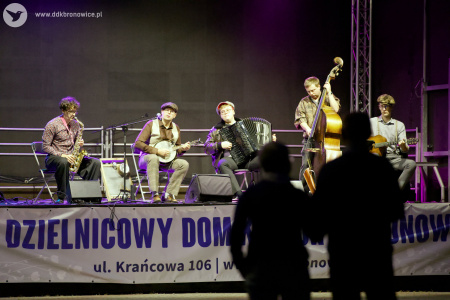 Na scenie, wieczorem koncert kapeli. Muzycy grają, na saksofonie, bandżo, kontrabasie, gitarze i akordeonie. Przed sceną kontury dwóch słuchających osób.