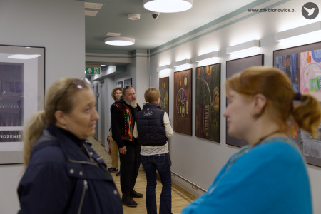 Młode osoby oglądają zawieszone obrazy w Galerii Przechodniej. Rozmawiają ze sobą i kontemplują sztukę. W tle Grzegorz Nowicki rozmawia z kobietą przy obrazach.