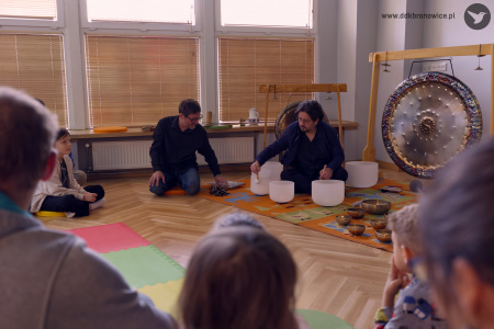 Dwóch artystów ubranych na czarno. Artysta po lewej stronie czyta książę. Artysta po prawej gra na białych misach.