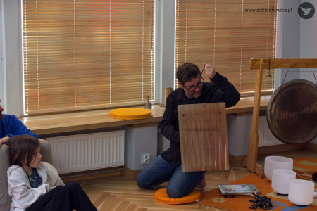 Artysta w czarnej koszuli wyciągnął długi instrument zawieszony na sznurkach. Wygląda jak cymbałki.
