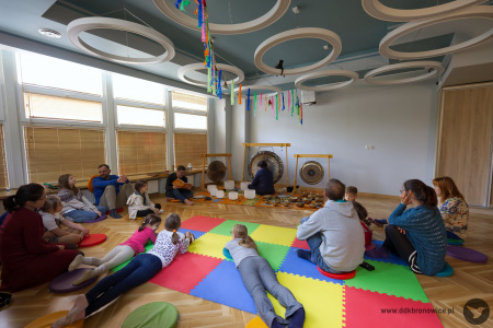 Sala Twórczej Pracowni. Uczestnicy siedzą na podłodze. Dwóch artystów ubranych na czarno siedzi wokół mis i gongów. Z sufitu zwisają kolorowe wstążki.