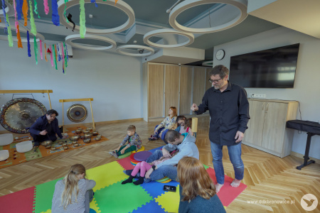 Artysta w czarnej koszuli chodzi między uczestnikami. Uczestnicy mają zamknięte oczy. Artysta porusza drewnianym, delikatnym instrumentem.
