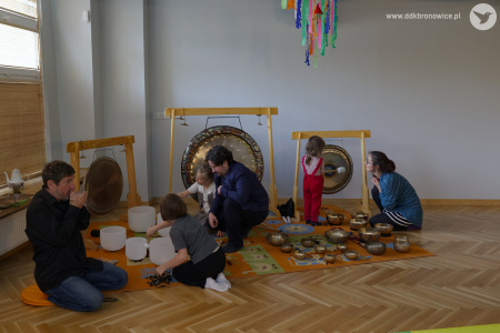 Wokół artystów siedzą dzieci. Uczą się grać na instrumentach. Artyści im pomagają. Dzieci mogą dotknąć instrumentów lub zagrać dźwięki.