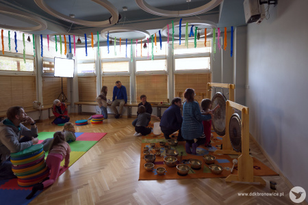 Sala Twórczej Pracowni. Wokół artystów siedzą dzieci. Uczą się grać na instrumentach. Artyści im pomagają. Dzieci mogą dotknąć instrumentów lub zagrać dźwięki.