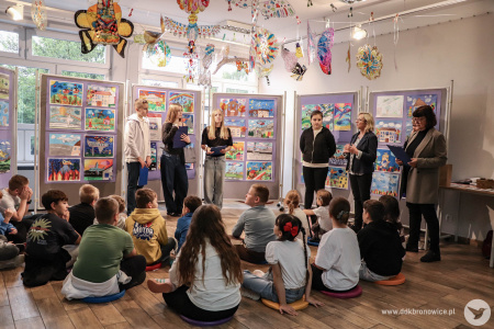 Galeria Mała. Uczniowie i uczennice siedzą na kolorowych poduszkach. Trójka starszych uczniów prowadzą wydarzenie ze scenariuszami w rękach. Po prawej stronie dwójka nauczycieli i Ewa Rejmak.