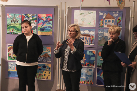 Koordynatorka Galerią Małą, Ewa Rejmak, wygłasza przemówienie do zgromadzonych uczniów i uczennic. Za nią zawieszone prace.