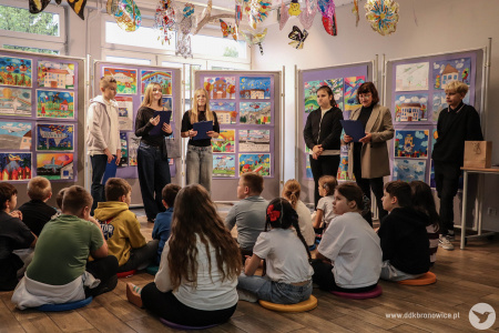 Galeria Mała. Uczniowie i uczennice siedzą na kolorowych poduszkach. Trójka starszych uczniów prowadzą wydarzenie ze scenariuszami w rękach. Po prawej stronie dwójka nauczycieli i Ewa Rejmak.