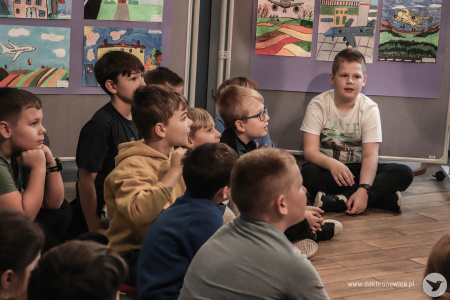 Uczniowie i uczennice siedzą i słuchają uważnie prowadzących. Siedzą na kolorowych poduszkach. Za nimi stoją zawieszone prace.