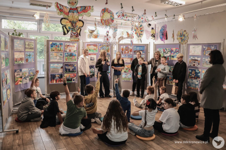 Galeria Mała. Uczniowie i uczennice siedzą na kolorowych poduszkach. Trójka starszych uczniów prowadzą wydarzenie ze scenariuszami w rękach. Po prawej stronie dwójka nauczycieli, Ewa Rejmak i Aleksandra Szulc-Choma.