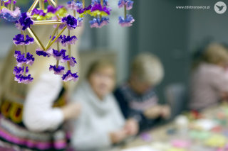 Grafika promująca wydarzenie cykliczne Na Ludowo. Grafikę stanowi beżowy materiał ze słowiańskim wzorem. W środkowej części grafiki zdjęcie słomy, na której zawieszone są kolorowe kwiaty. Obok nich znajdują się zapalone lampki i beżowy obrus. Na samej górze grafiki, brązowo-czerwony napis: „NA LUDOWO”, na białym tle. Wokół napisów czerwone koraliki. Po lewej stronie grafiki, błękitny napis: „SŁOMIANE/OZDOBY”. W lewym dolnym rogu, na beżowym tle, biały napis: „WARSZTATY W RAMACH FESTIWALU BOŻEGO NARODZENIA”. W prawym dolnym rogu napis: www.ddkbronowice.pl, obok napisu mały koliber na szarym tle.