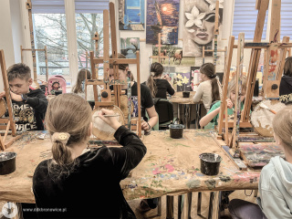 Grafika promująca program zimowych warsztatów artystycznych w Dzielnicowym Domu Kultury “Bronowice”. Na granatowym tle widać padający śnieg. Na dole grafiki znajduje się bałwanek. Ubrany jest w niebieski szalik, buty, rękawiczki i nauszniki. Stoi na śniegu, na jednej nodze, drugą uniesioną ma w górze. Ręce ma skierowane do góry. Ma zamknięte oczy i szeroki uśmiech. Po prawej stronie grafiki napis: “ZIMA Z KULTURĄ”, pod nim: “DZIELNICOWY DOM KULTURY “BRONOWICE””. W prawym dolnym rogu szary napis: www.ddkbronowice.pl, obok napisu mały koliber na szarym tle.