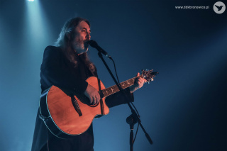 Grafika promująca koncert Jana Kondraka w Pracowniach Kultury Maki. Po lewej stronie grafiki postać Jana Kondraka w czarnej koszuli na jasnym tle. W prawym górnym rogu pomarańczowe napisy, od góry: “STACHURA/COHEN/OKUDŻAWA”. Pod napisami, czarny autograf Jana Kondraka. W prawym dolnym rogu szary napis: www.ddkbronowice.pl, obok napisu mały koliber na szarym tle.