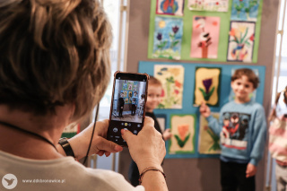 Grafika promująca wystawę prac dzieci z Przedszkola numer 7 w Pracowniach Kultury Maki. Grafikę stanowi obraz jednego z dzieci. Na białym tle kolorowe kwiaty z zielonymi łodygami. W lewym górnym rogu logo Galerii Małej: białe kontury postaci uśmiechniętego dziecka na czerwonym tle. Pod nim podpis: Galeria Mała. W prawym dolnym rogu napis www.ddkbronowice.pl, obok napisu mały koliber na szarym tle.